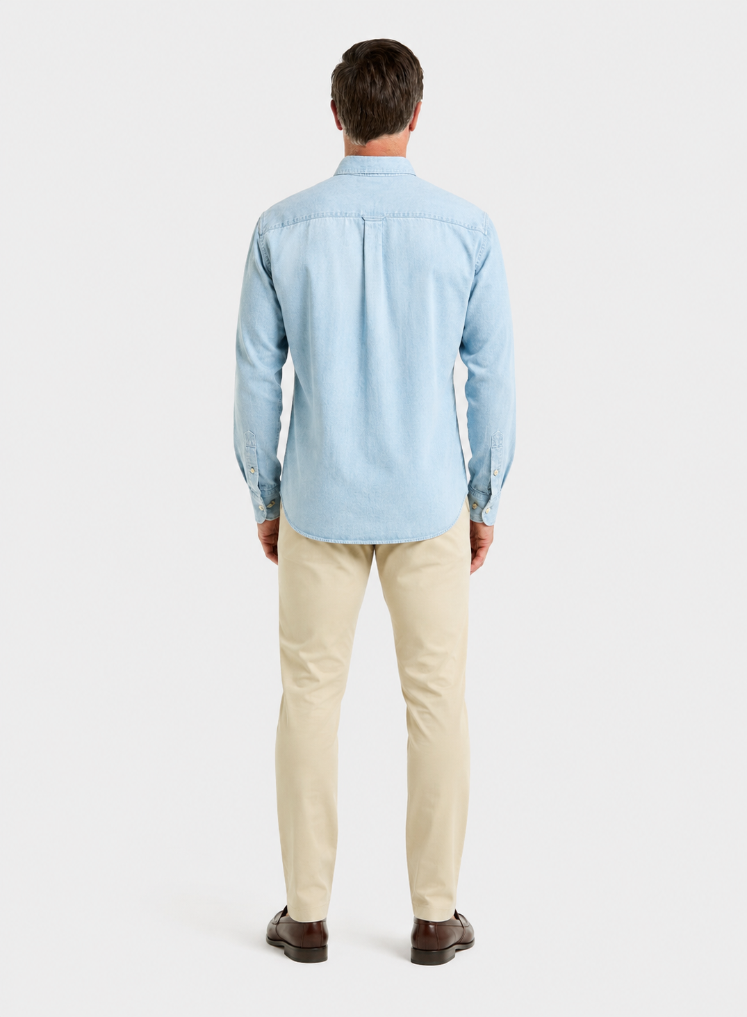 A man viewed from the back wearing a light blue long-sleeve button-up shirt, beige trousers, and dark brown shoes, standing against a light gray studio background.