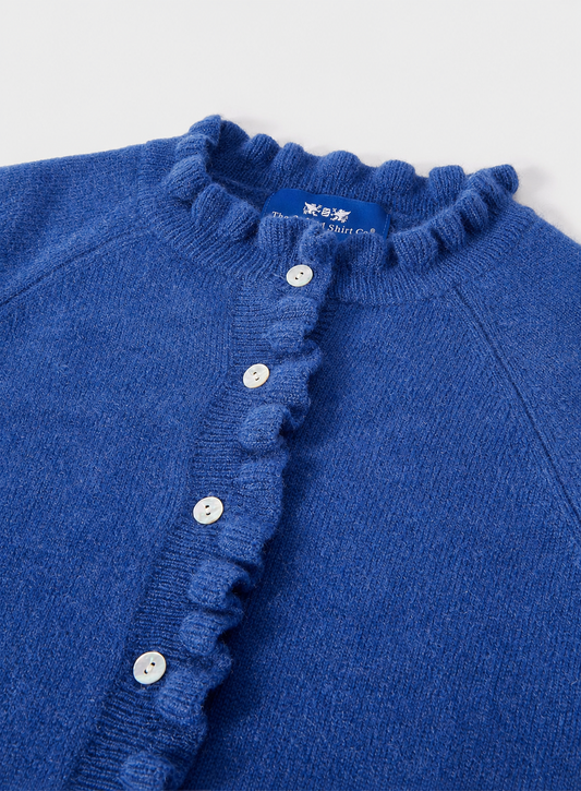 A close-up of a blue knit cardigan sweater featuring a ruffled collar and a matching ruffled button placket with white buttons, shown against a light gray background.