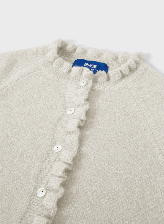 Close-up of a cream-colored knit cardigan featuring a ruffled collar and a ruffled button placket with white buttons. A blue brand label is visible at the neckline.