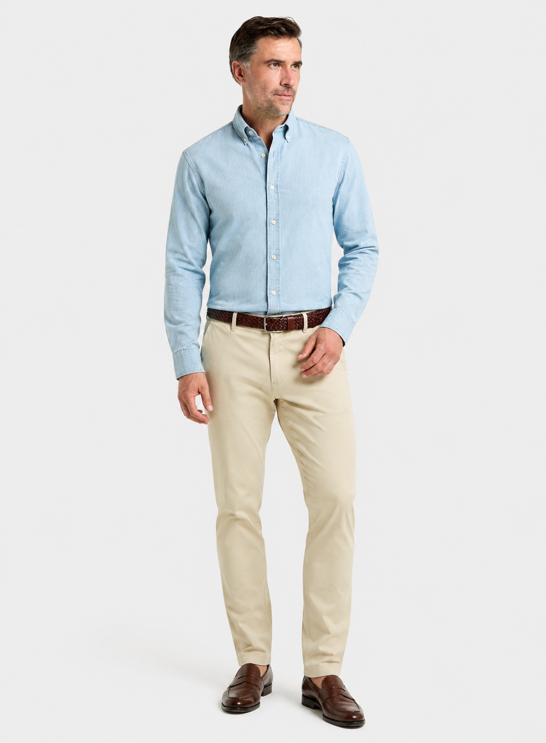 A man modeling a light blue long-sleeve button-down shirt, beige trousers, a brown belt, and brown leather dress shoes against a light gray studio background.