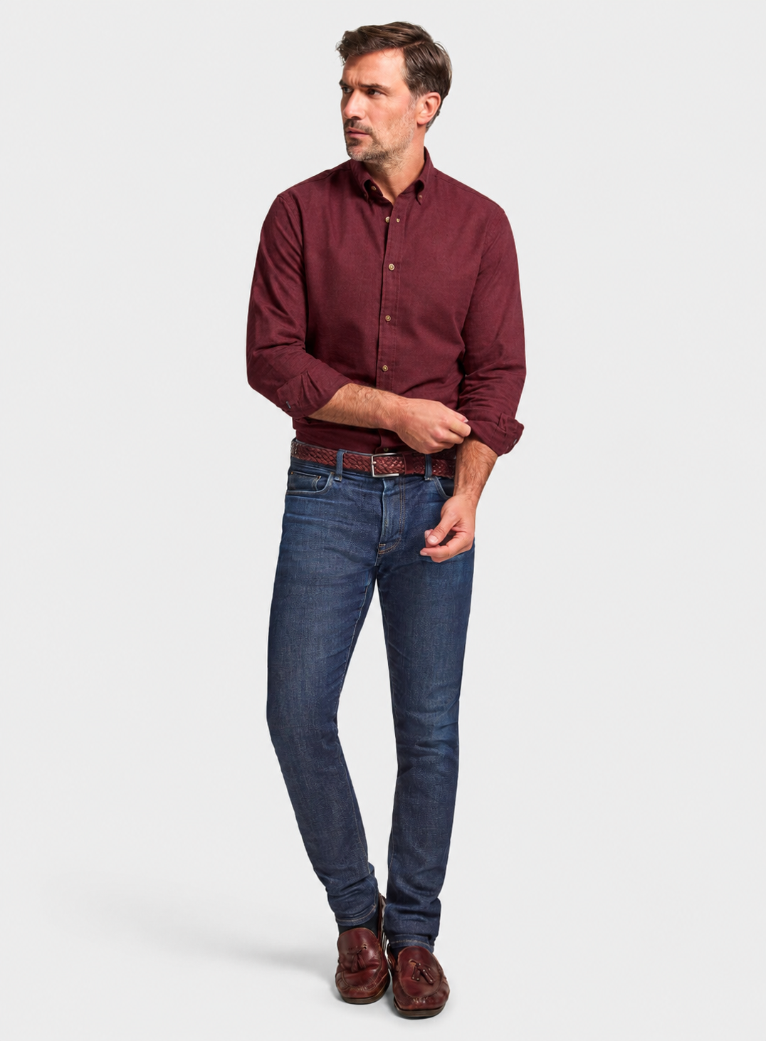 A man modeling a long-sleeve burgundy button-down shirt, dark blue jeans, a brown belt, and brown leather shoes against a light gray studio background.