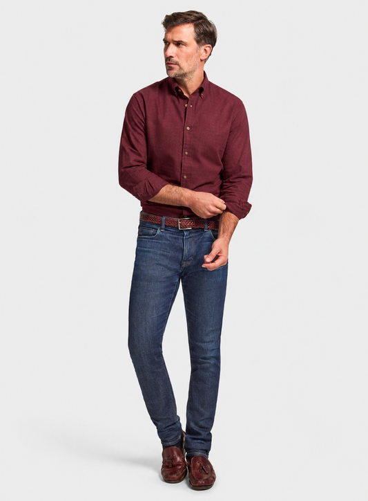 A man modeling a long-sleeve burgundy button-down shirt, dark blue jeans, a brown belt, and brown leather shoes against a light gray studio background.