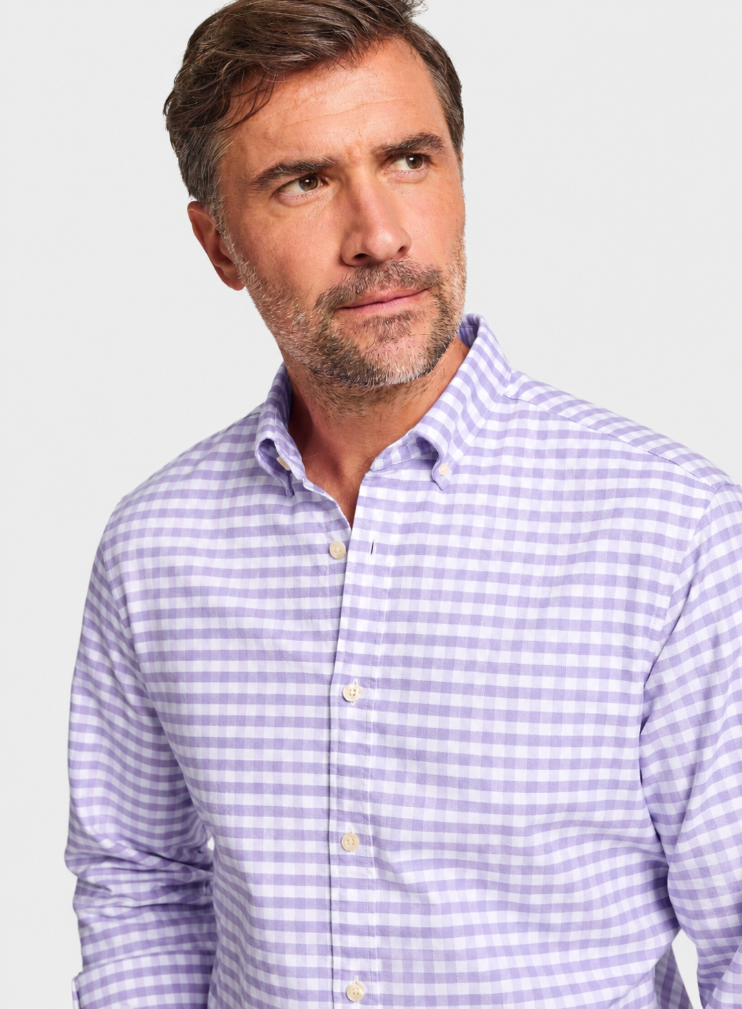 A man wearing a light purple and white gingham button-down shirt, shown from the chest up against a plain light gray studio background.