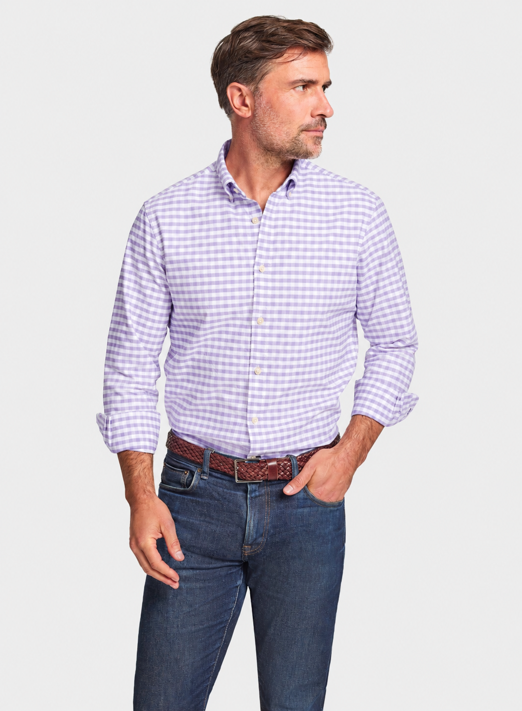 A man wearing a light purple and white gingham long-sleeve button-down shirt, paired with dark blue jeans and a brown belt, standing against a light gray background.