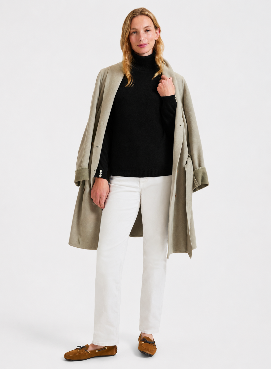 A woman modeling a light beige trench coat over a black roll neck sweater, white pants, and brown loafers, photographed against a plain light gray studio background.
