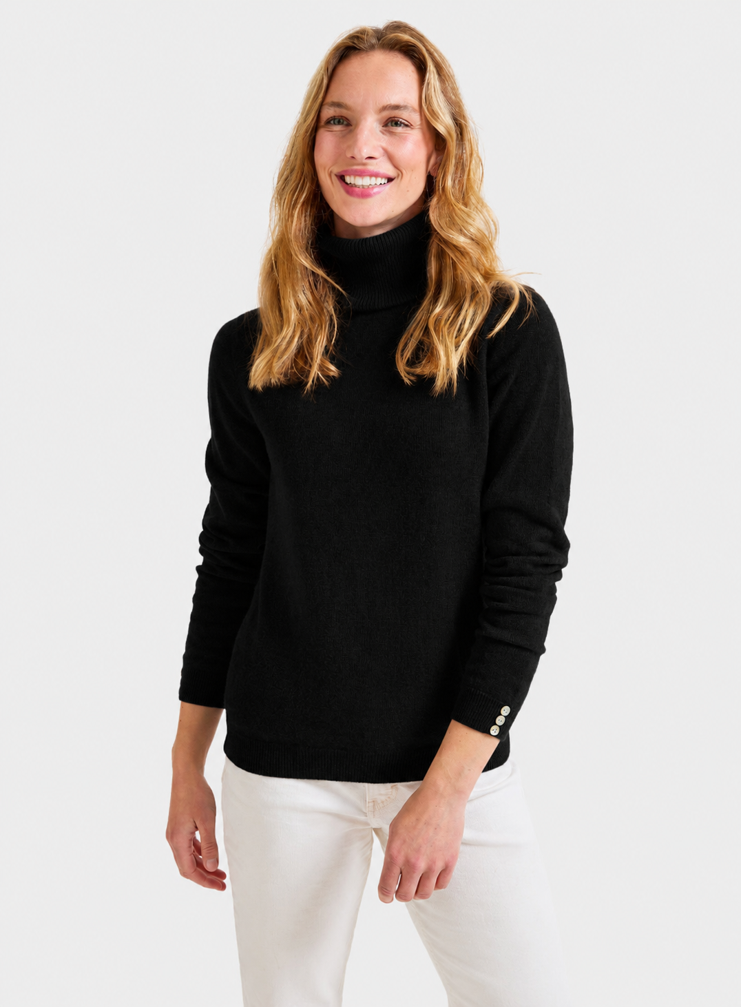 A woman wearing a black ribbed roll neck sweater and white pants, standing against a light gray studio background.