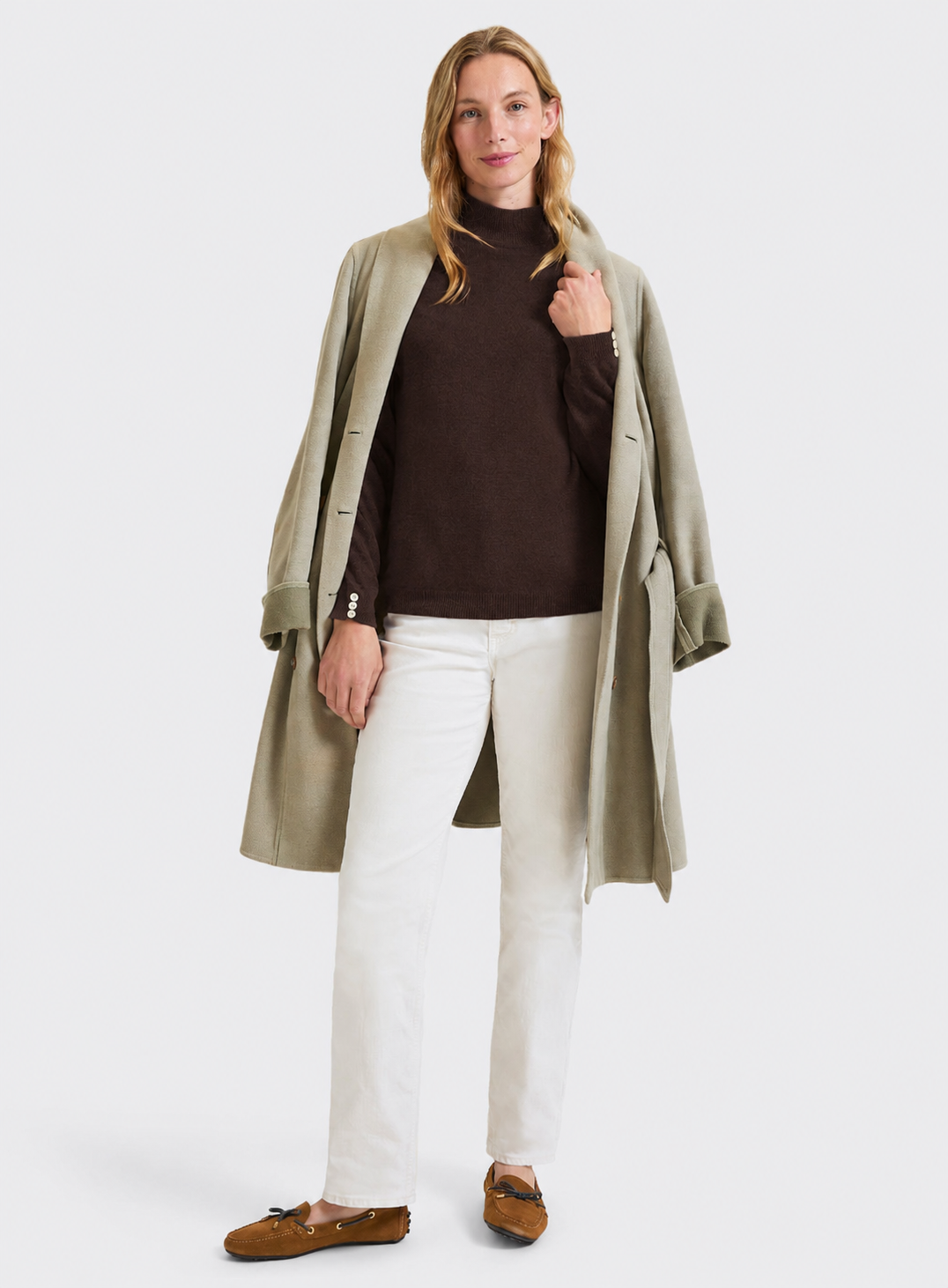 A woman modeling a light olive green mid-length coat over a dark brown turtleneck sweater, white pants, and brown loafers, photographed against a solid light gray studio background.