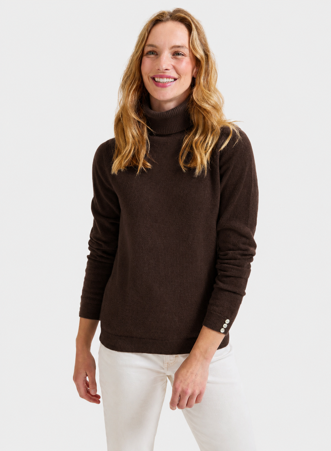 A woman with blonde hair wearing a dark brown ribbed turtleneck sweater and white pants, standing against a light gray studio background.