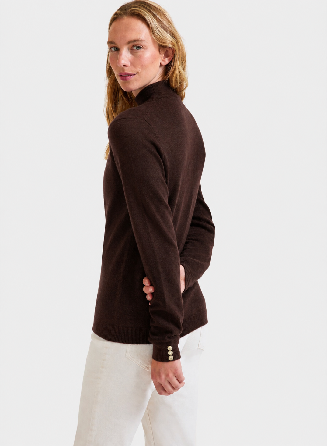 A woman wearing a dark brown mock-neck long-sleeve knit top with decorative gold buttons on the cuffs, paired with white pants, photographed against a light gray studio background.