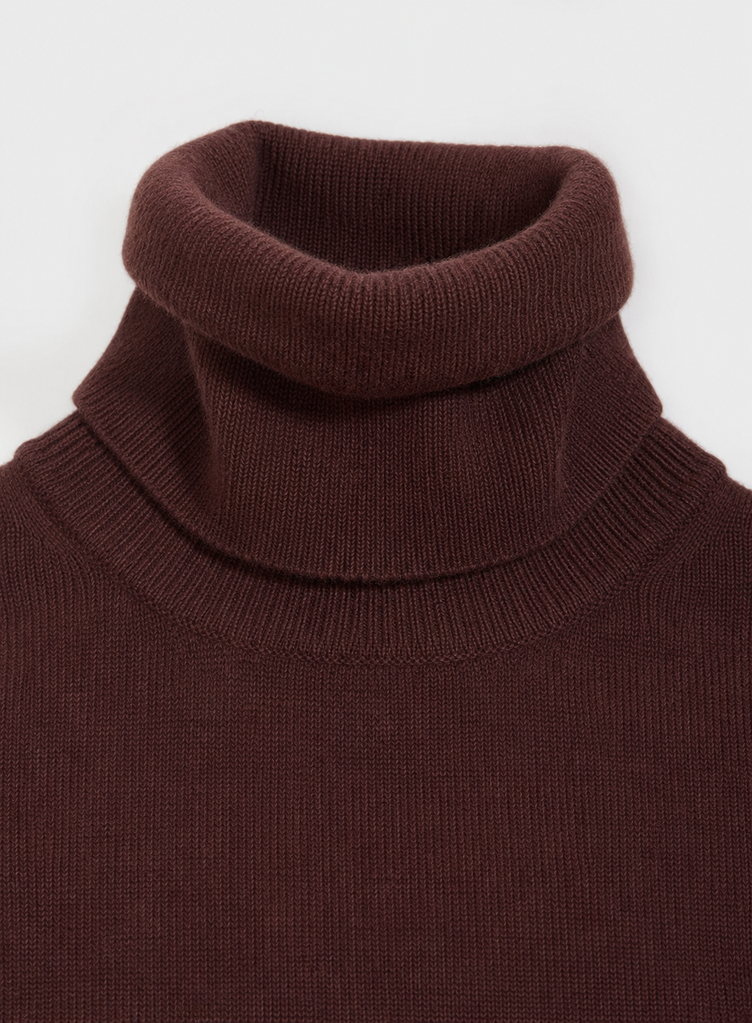 Close-up of a ribbed knit roll neck sweater in a deep dark brown color, showing the texture and collar detail against a light gray background.
