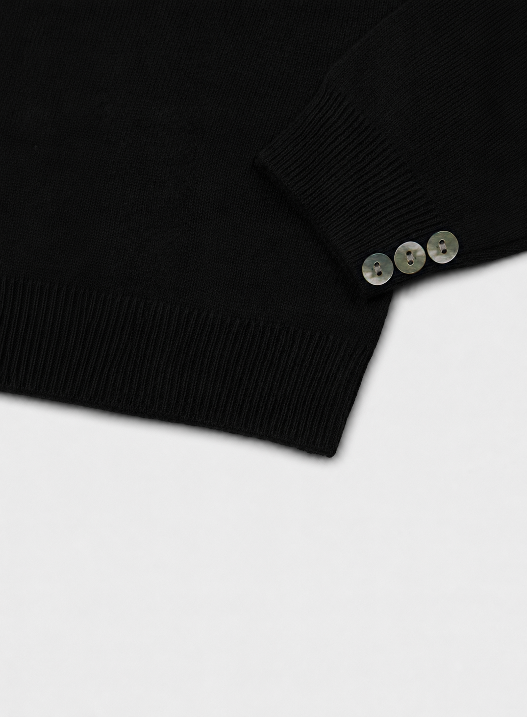 Close-up of a black ribbed knit garment, cashmere roll neck, with three small metallic buttons near the cuff on a light gray background.