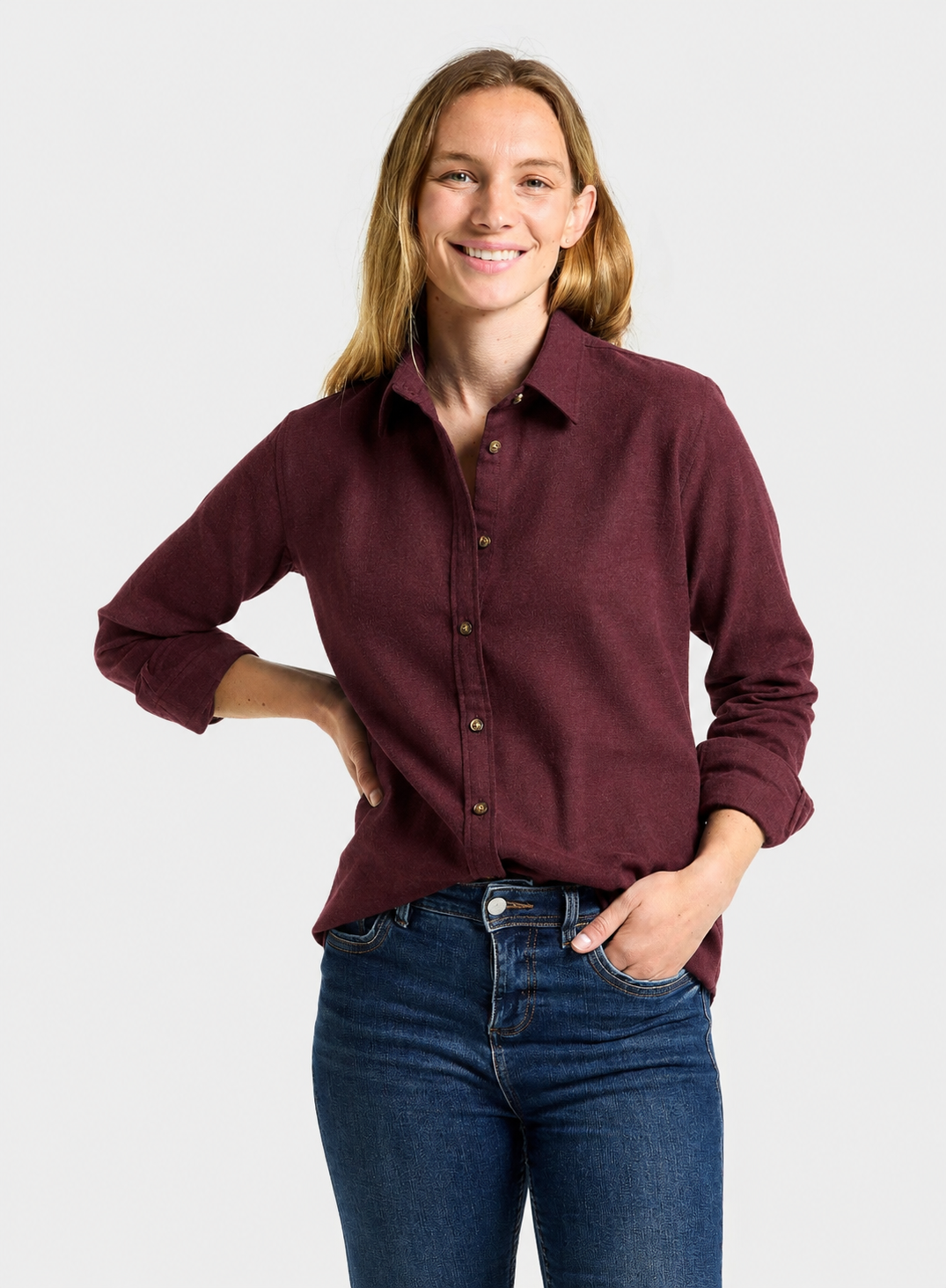 A woman wearing a long-sleeve button-up shirt in maroon and blue jeans, standing against a light gray studio background.