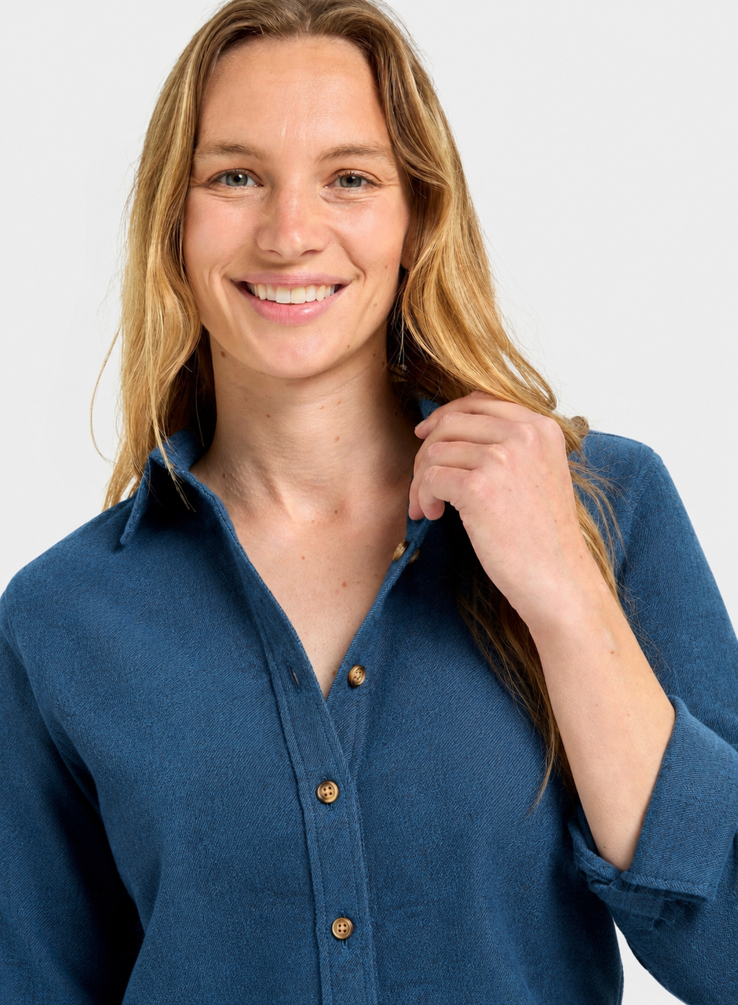 A smiling blonde woman wearing a blue button-up shirt with rolled sleeves and brown buttons, photographed against a light gray studio background.