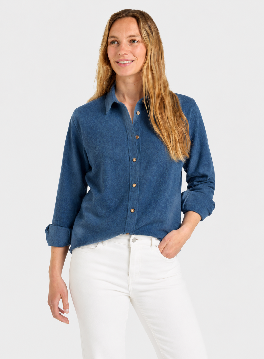 A woman modeling a blue long-sleeve button-up shirt with white pants against a light gray studio background.