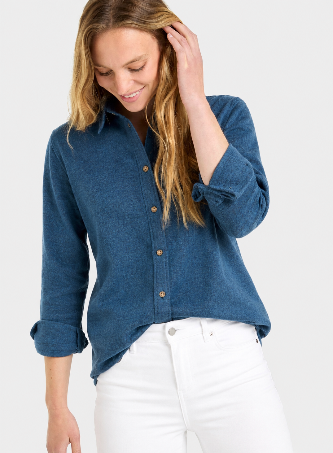 A woman wearing a blue long-sleeve button-up shirt with rolled cuffs and white pants, photographed against a light gray studio background.