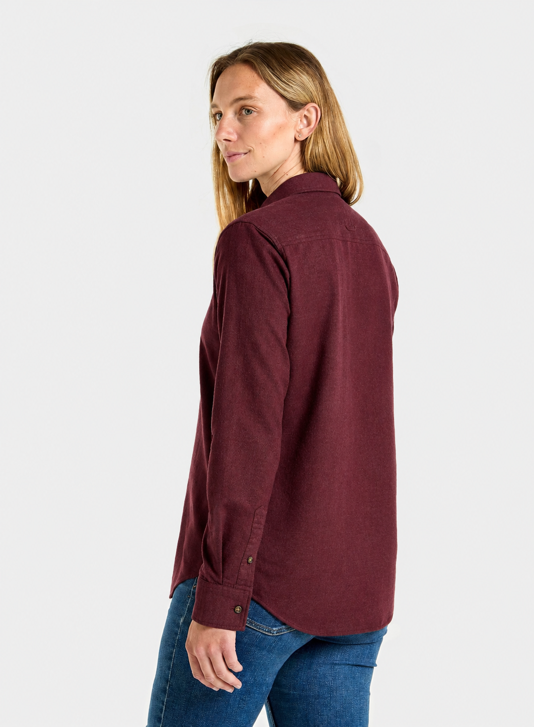 A woman shown from the side-back wearing a long-sleeve button-up shirt in deep burgundy with blue jeans, photographed against a plain light gray studio background.