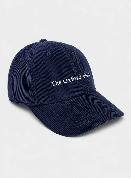 A navy blue corduroy baseball cap with 'The Oxford Shirt' embroidered in white on the front, set against a light gray background.