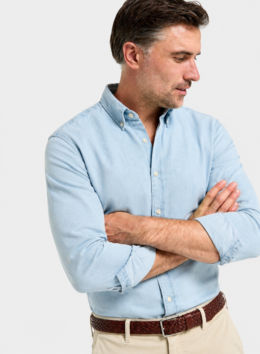 A man wearing a light blue long-sleeve button-down shirt, paired with beige trousers and a brown woven belt, photographed against a plain light gray studio background.