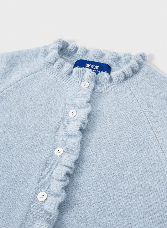 A close-up of a light blue knit cardigan sweater featuring a ruffled collar and a ruffled button placket with white buttons. A blue brand label is visible inside the collar.