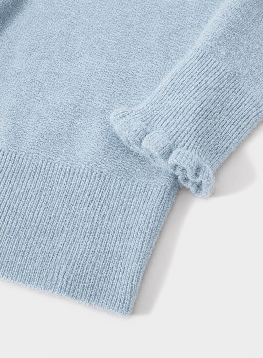 Close-up of a light blue knit garment showing a ribbed cuff with a decorative ruffled edge, on a plain light background.