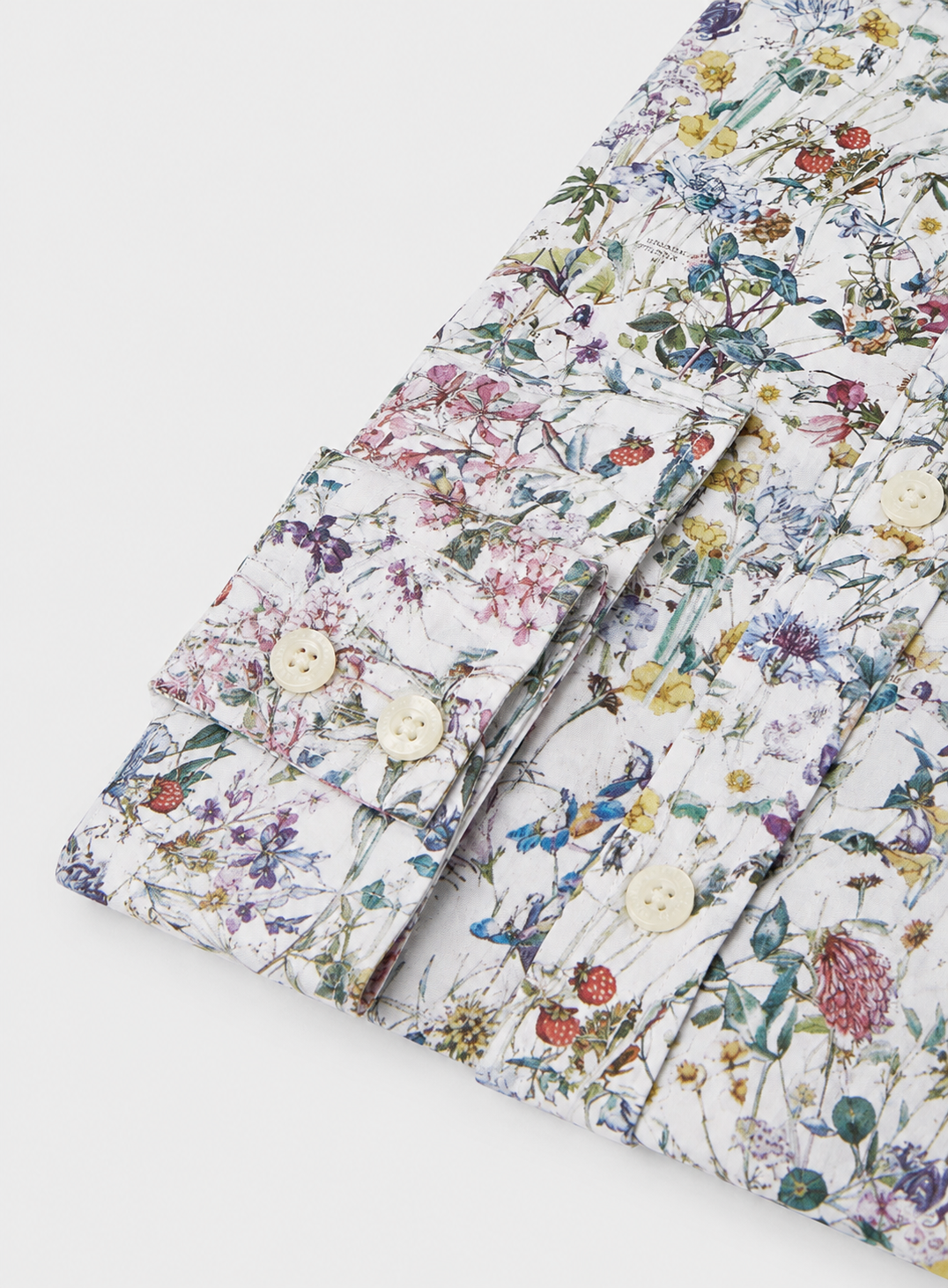 A close-up of a folded long-sleeve button-up shirt with a dense, multicolor wildflower and bird print on a white background, featuring light beige buttons.