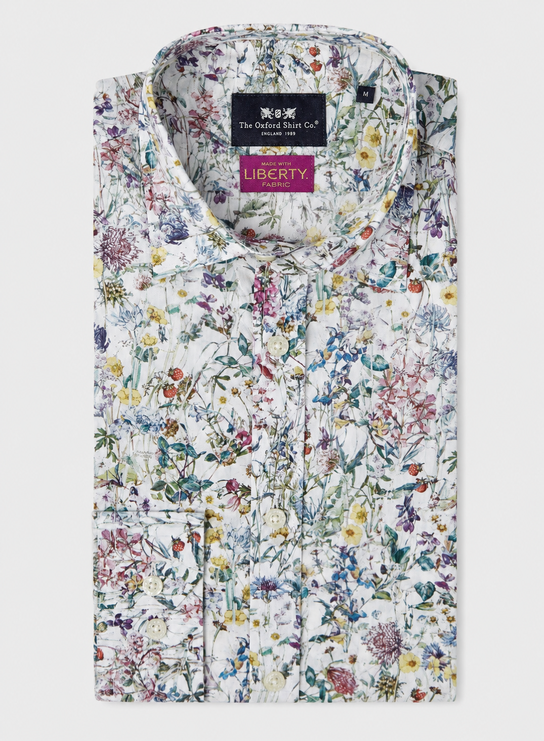 A folded white cotton shirt with a dense multicolor floral print, featuring a black and pink Liberty brand label on the collar, set against a light gray background.