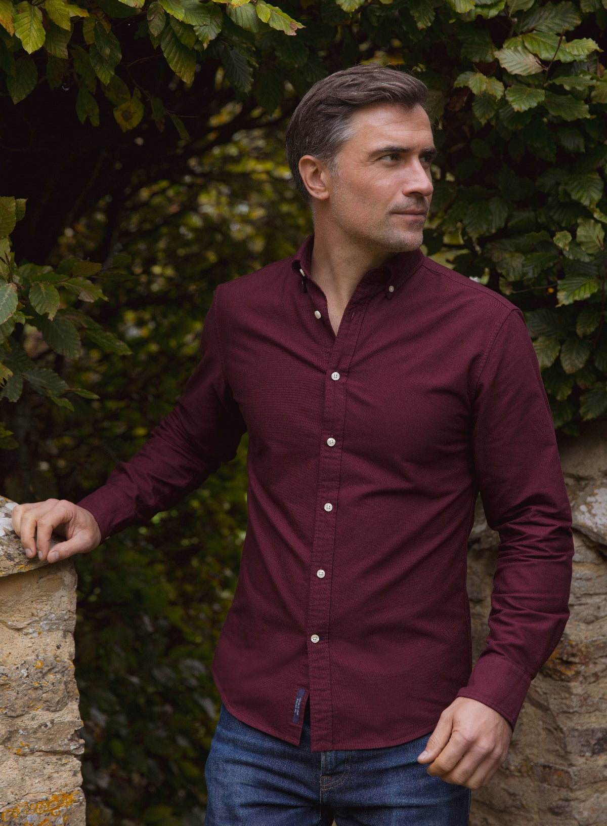 A man wearing a long-sleeve burgundy button-down shirt and blue jeans, standing outdoors against a background of green foliage and a stone wall.