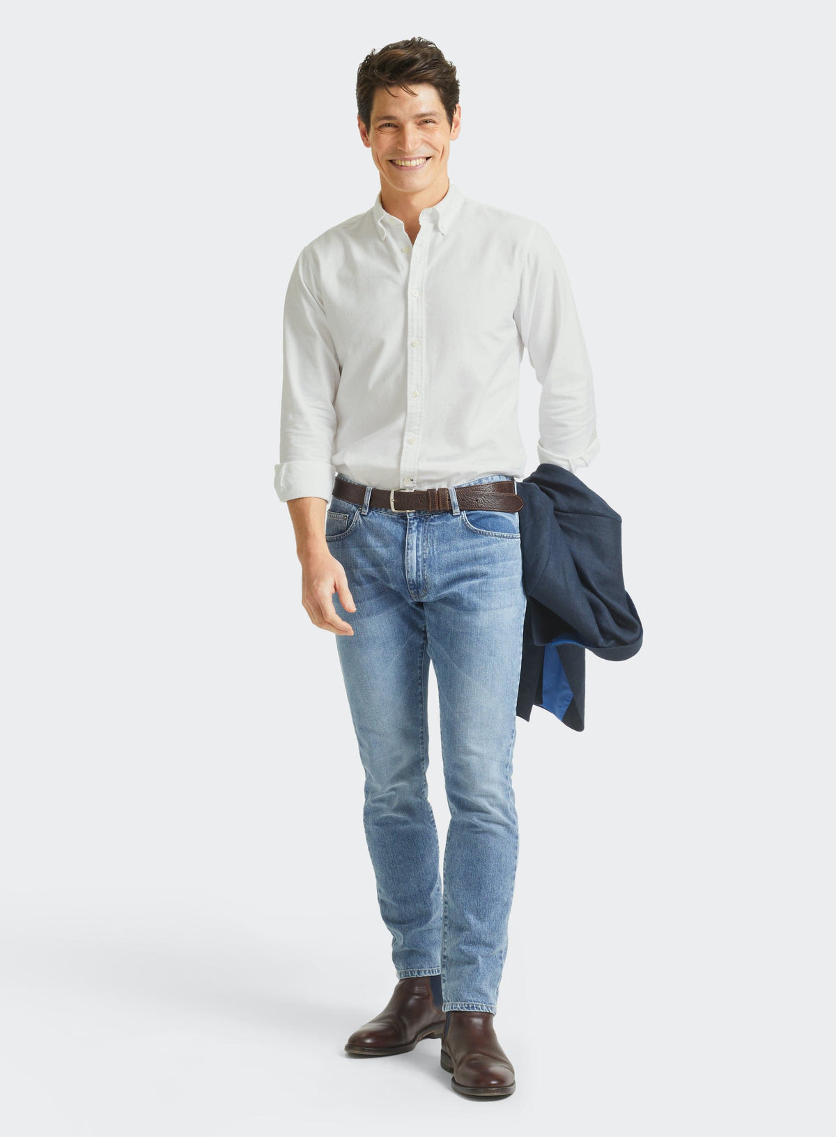 Man wearing a men's white Oxford button-down shirt with blue jeans, holding a navy jacket